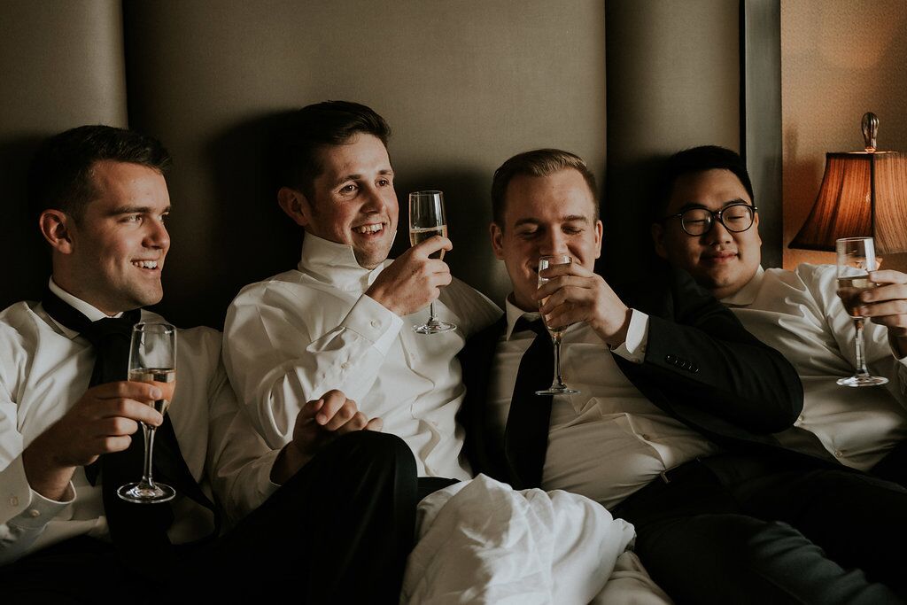 Groom and groomsman raise a glass all crowded onto bed at Oak Bay Beach HOtel