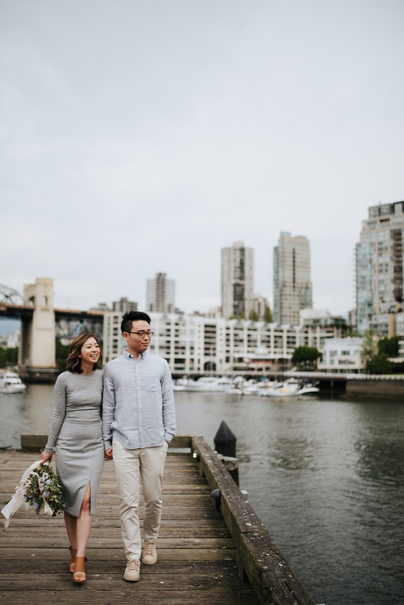 Vancouver Cityscape behind engaged couple who hold hands along the water