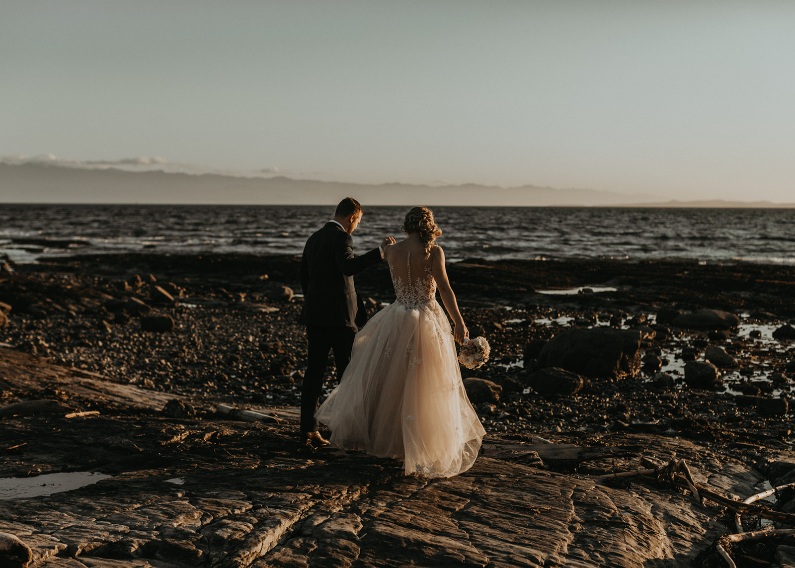 Bride and Groom walk along the rocky beach at reception