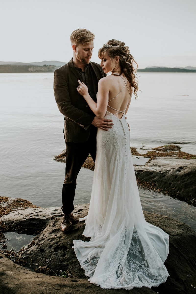 Bride in white lace gown with cutout back stands with groom in brown suit in front of the beach on Vancouver Island