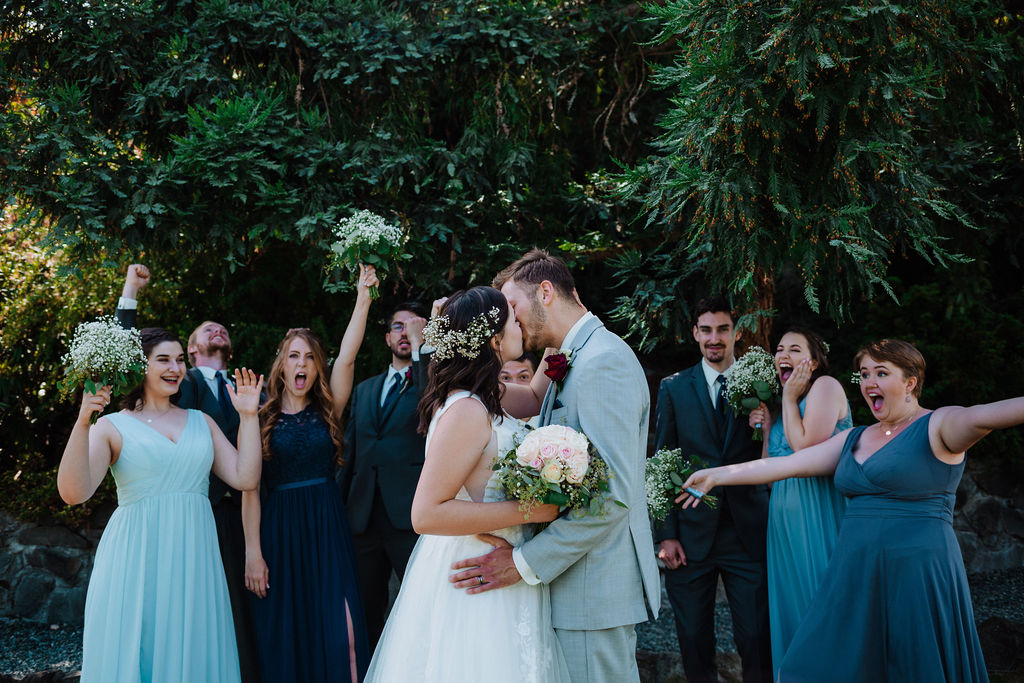 Newlyweds and bridal party laugh around the newlyweds kissing