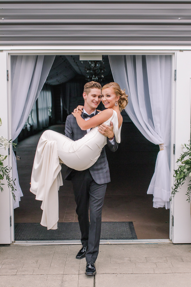 Groom in grey suit carries his bride in his arms at New Falls Golf Course