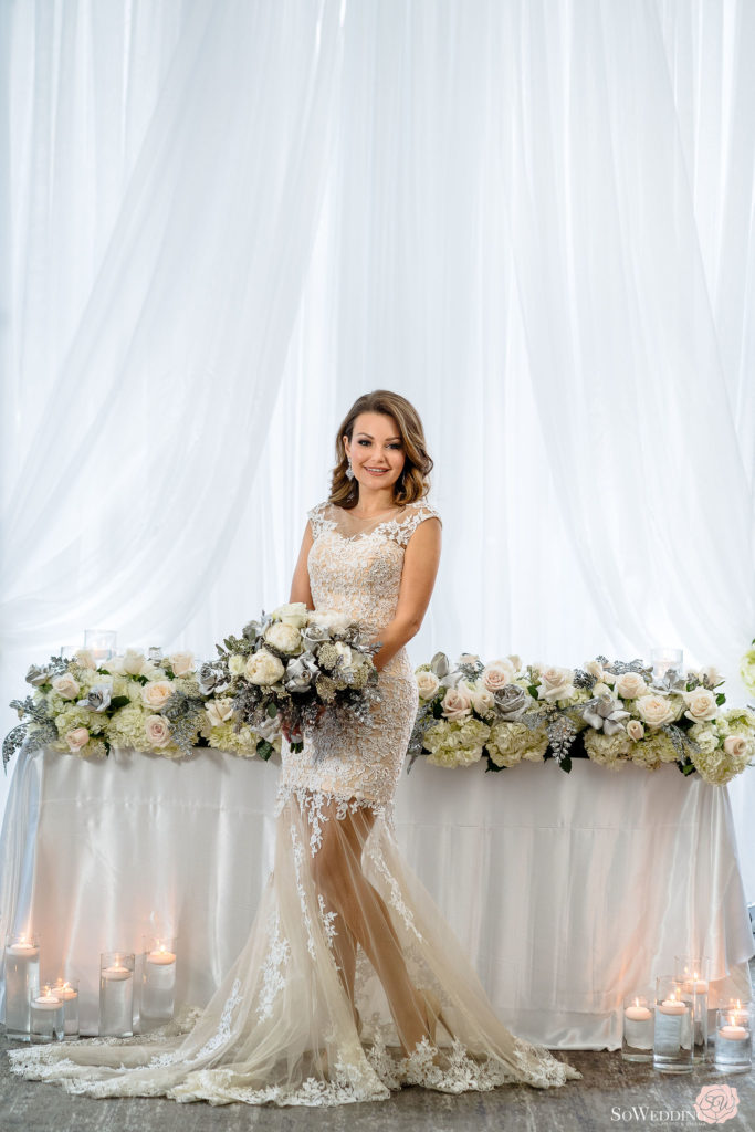 Bride holding silver, blue and white bouquet in front of head table with flower edge by Proline Trading Vancouver