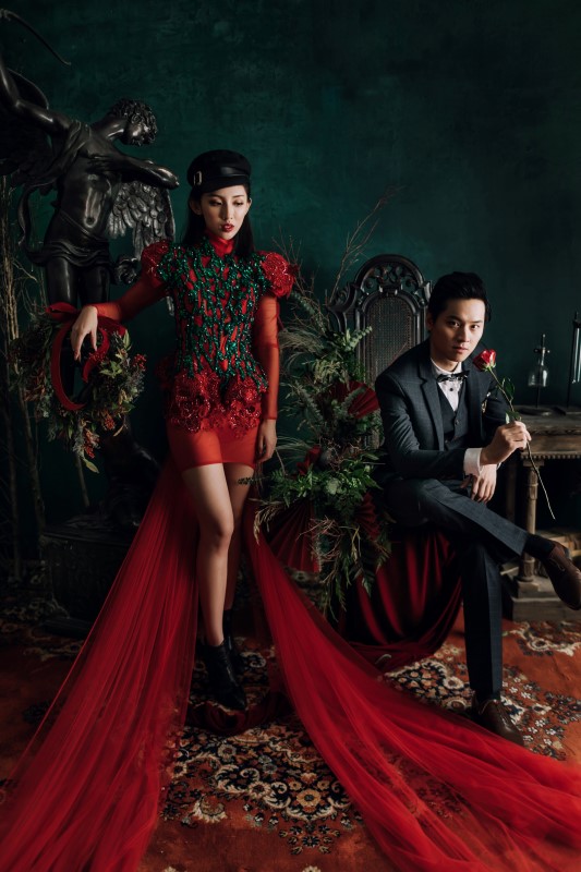 Culture Fusion bride in red gown with long train standing next to black angel statue, groom sitting on chair next to her