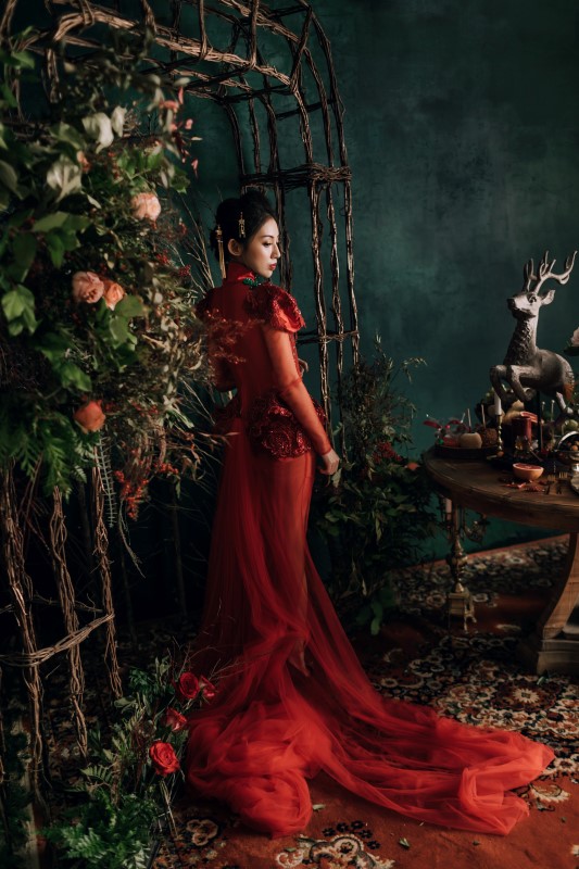 Culture Fusion bride in red gown with train standing next floral arrangements with orange roses