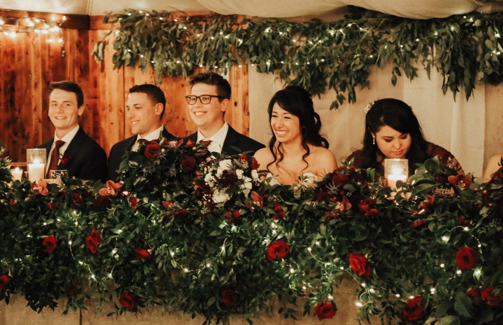 Steal of a Wedding head table with smiling bridal party and a lot of greenery, fairy lights and red roses