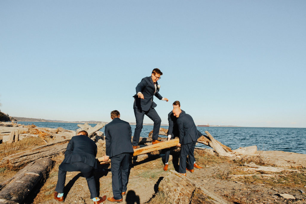 Steal of a Wedding groomsmen holding up a log at the beach with the groom standing on it