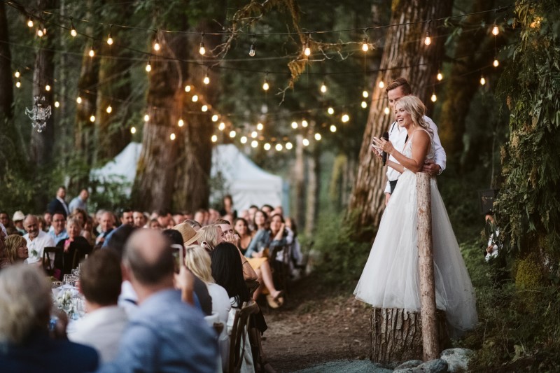 Bride and Groom stand on tree stump under globe lights giving speech to forest reception guests