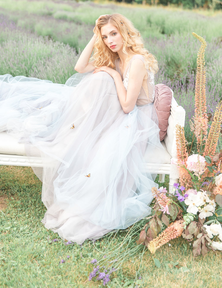 Bride Sits on Bench in LuxxNova gown surrounded by flowers in lavender field