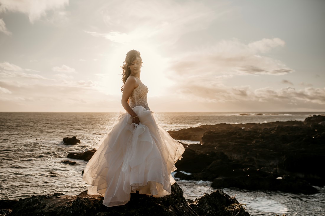 Bride stands on the black rock of Ucluelet holding her gown from Shades of White Vancouver Island