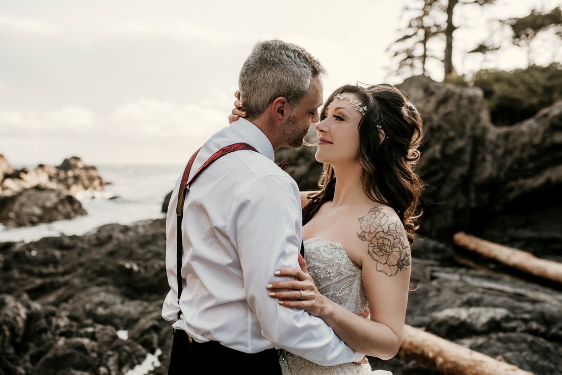 Bride with arm tattoo and groom wearing suspenders stand on beach surrounded by driftwood