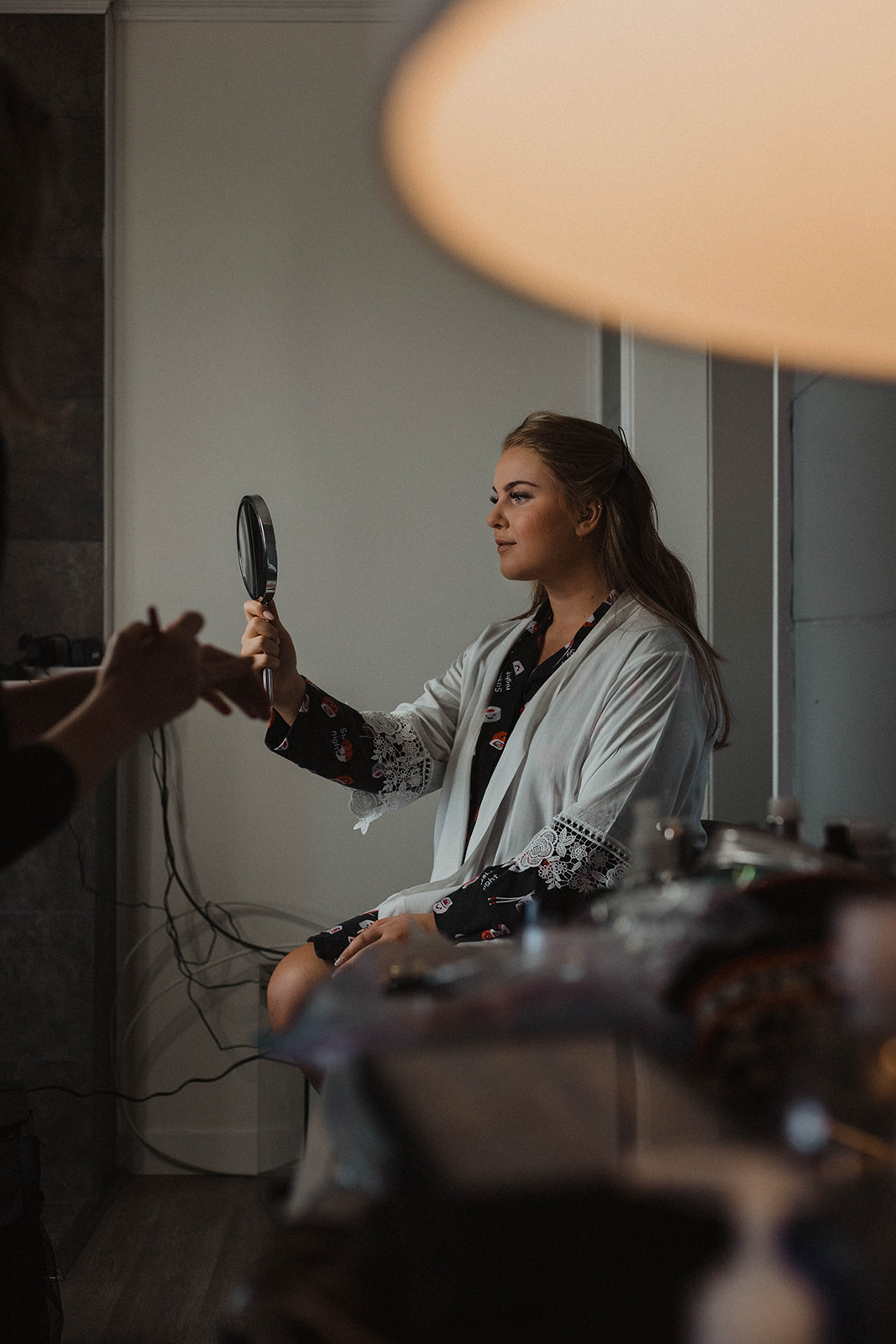 Bride getting hair done looks into hand mirror by Rory James