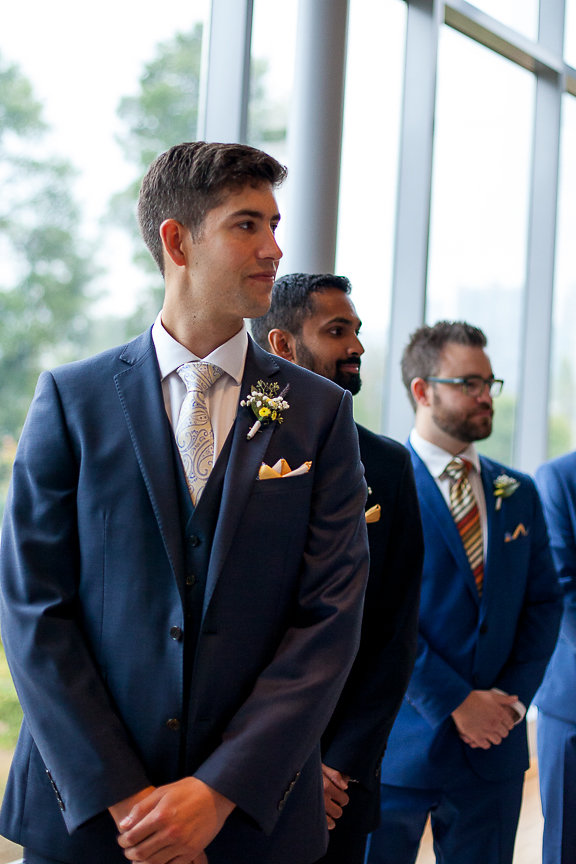 Groom in blue suit awaits bride at the end of aisle at Museum of Vancouver