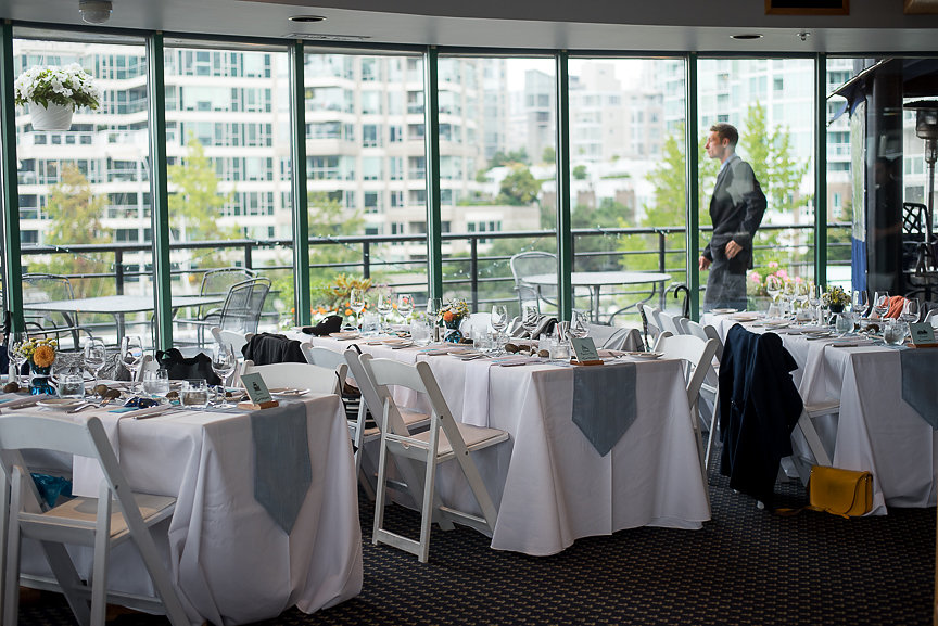 Guest tables in white linen and blue runners in front of bank of windows looking out at water False Creek Yacht Club Vancouver