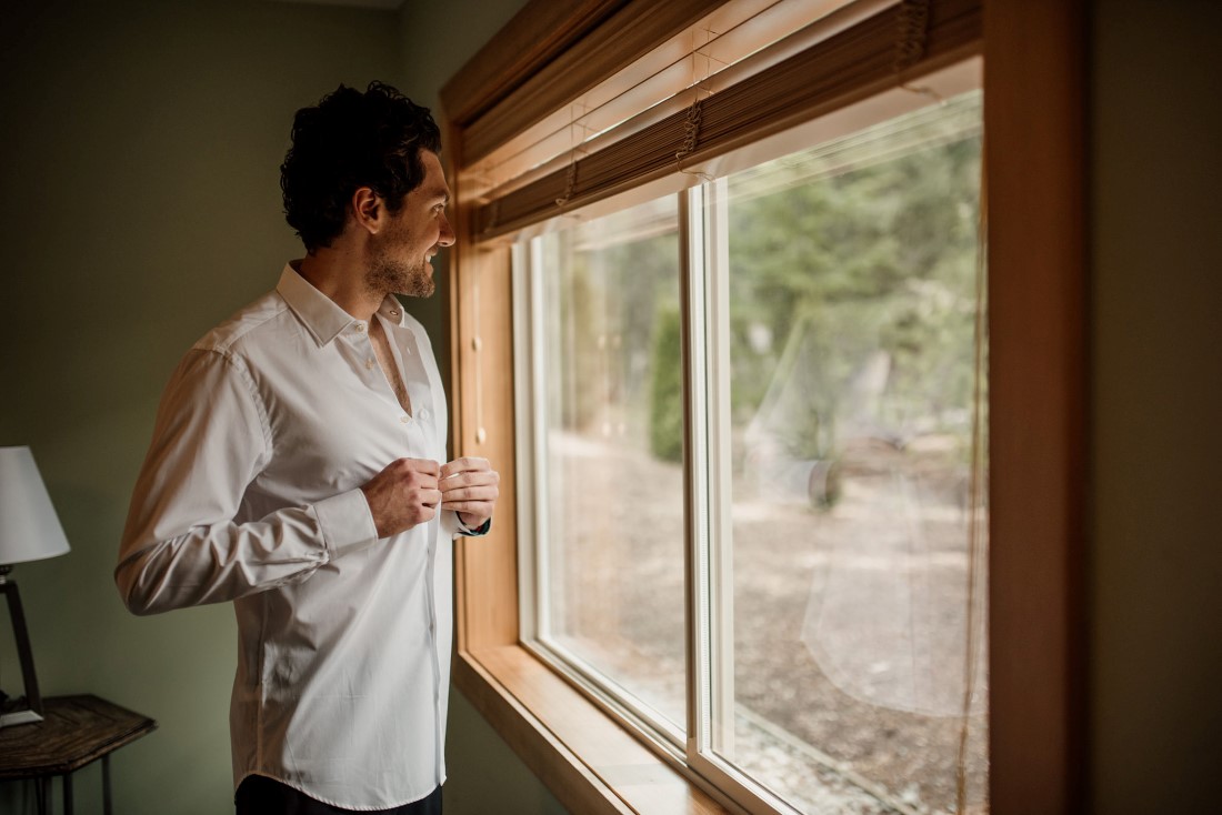 Groom buttons his shirt while looking out a window near Stone Circle Whistler