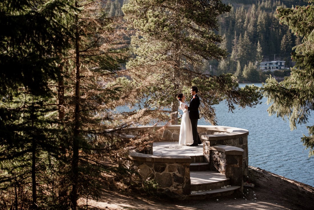 Stone Circle Whistler Wedding Couple stand among fir trees with lake behind 