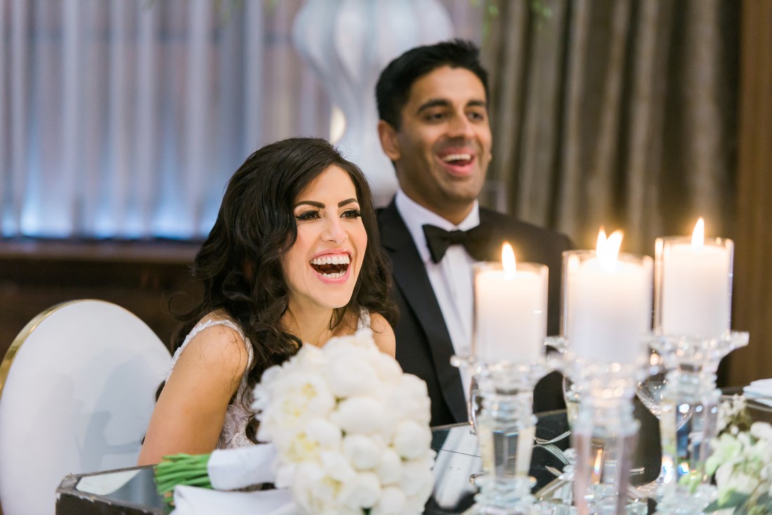 Bride and Groom laugh at speeches while seated at head table with candles and white roses at Vancouver Club 