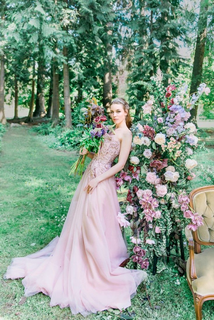 Bride wearing blush pink Majesty Bespoke wedding gown leans against flowers in Vancouver garden