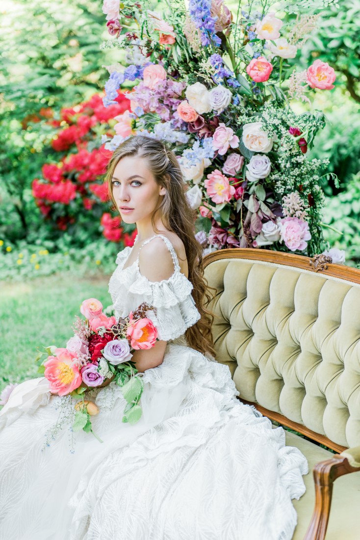 Bride in white lace flounce dress sits on settee in garden