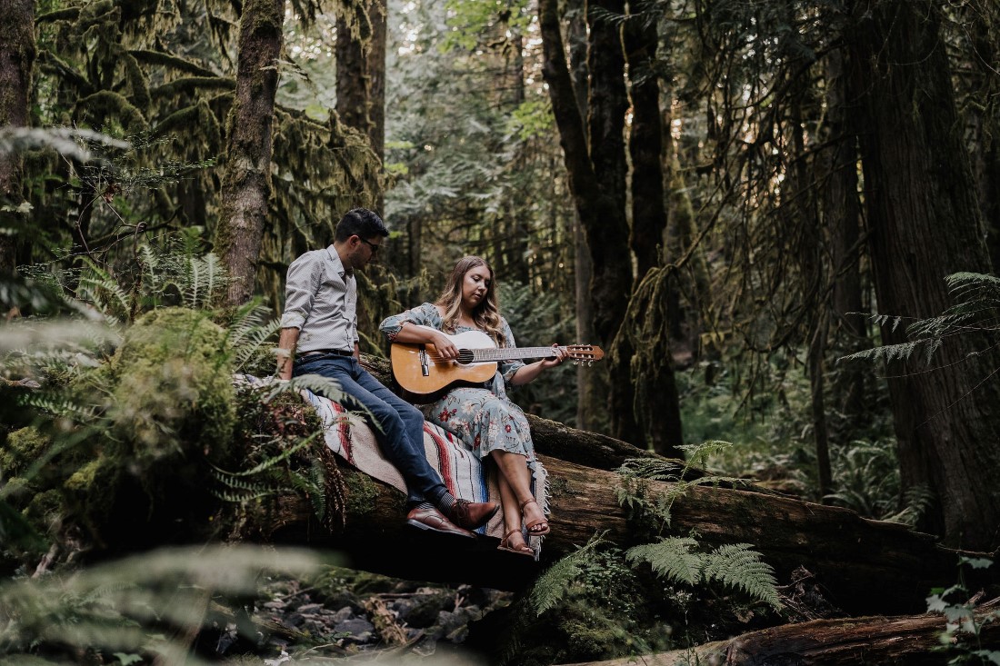 Engaged Couple sits on fallen moss covered tree and play guitar on Vancouver Island