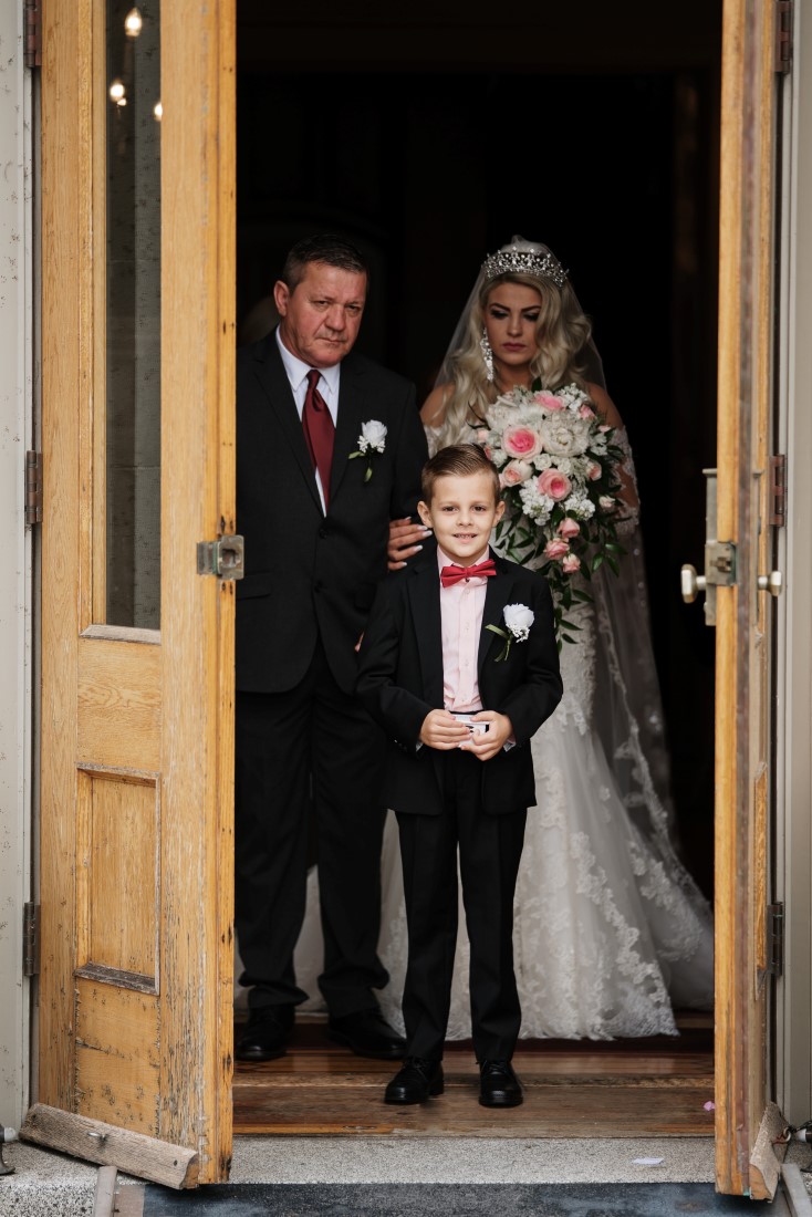 Castle Garden Wedding bride comes out of Hatley Castle doors with father and ringbearer