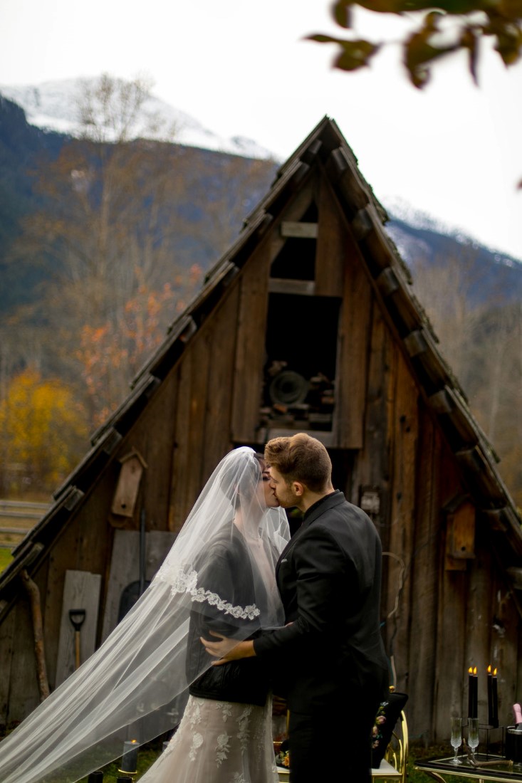 Bride and groom kiss in front of cabin and mountains wearing black leather jackets