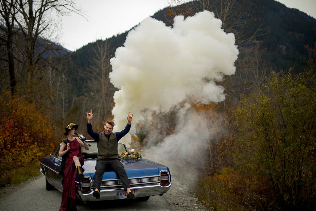 Smoke bomb behind blue classic car and newlyweds in BC mountains