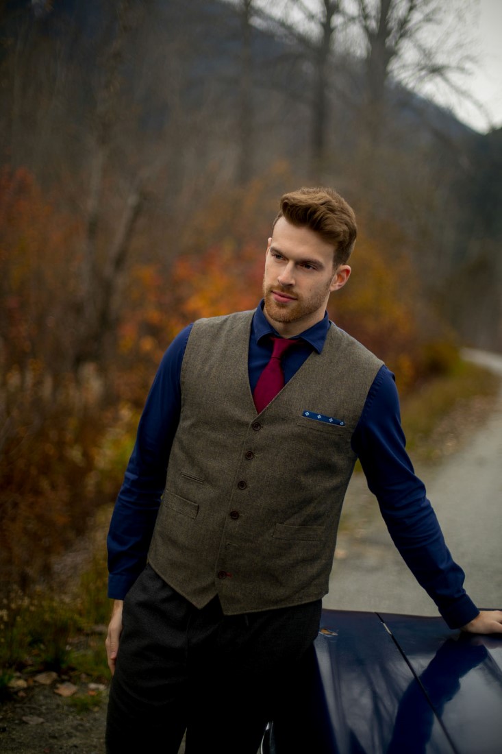 Groom leans on classic car wearing brown vest and burgundy tie