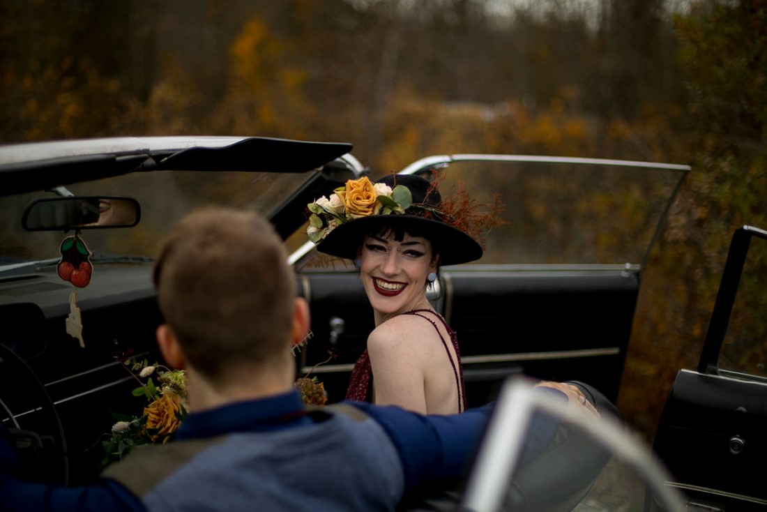 Bride smiles to camera wearing black hat with floral in classic car by Whisteria Events and Rentals