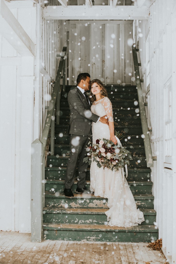 Snowy Urban Fairytale Wedding couple stand on stairs of barn with snowflakes all around