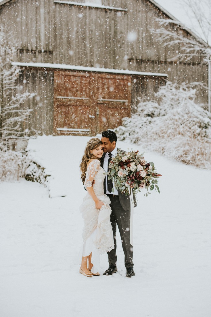 Bride and Groom stand in front of barn while large snowflakes fall on them