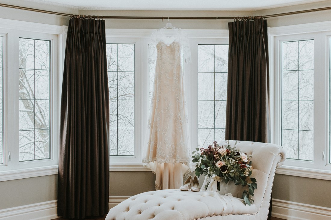 Maggie Sottero lace gown hangs in window while bouquet and shoes sit below on white lounge