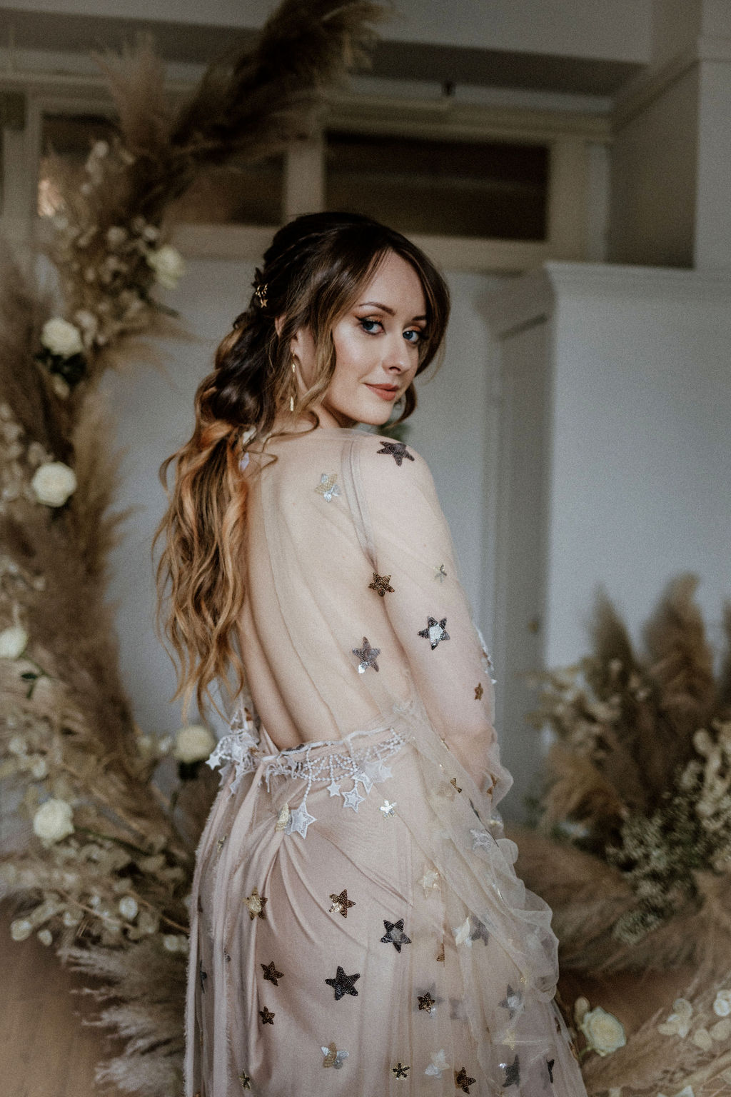 Bride wearing gold star gown by Flutters Dresses stands in front of pampas grass and white roses