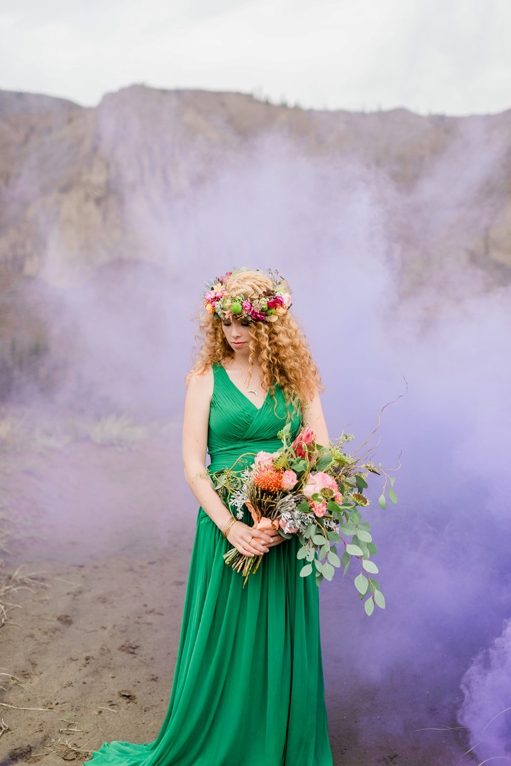 Bride in emerald green gown holds bouquet with purple smoke behind her