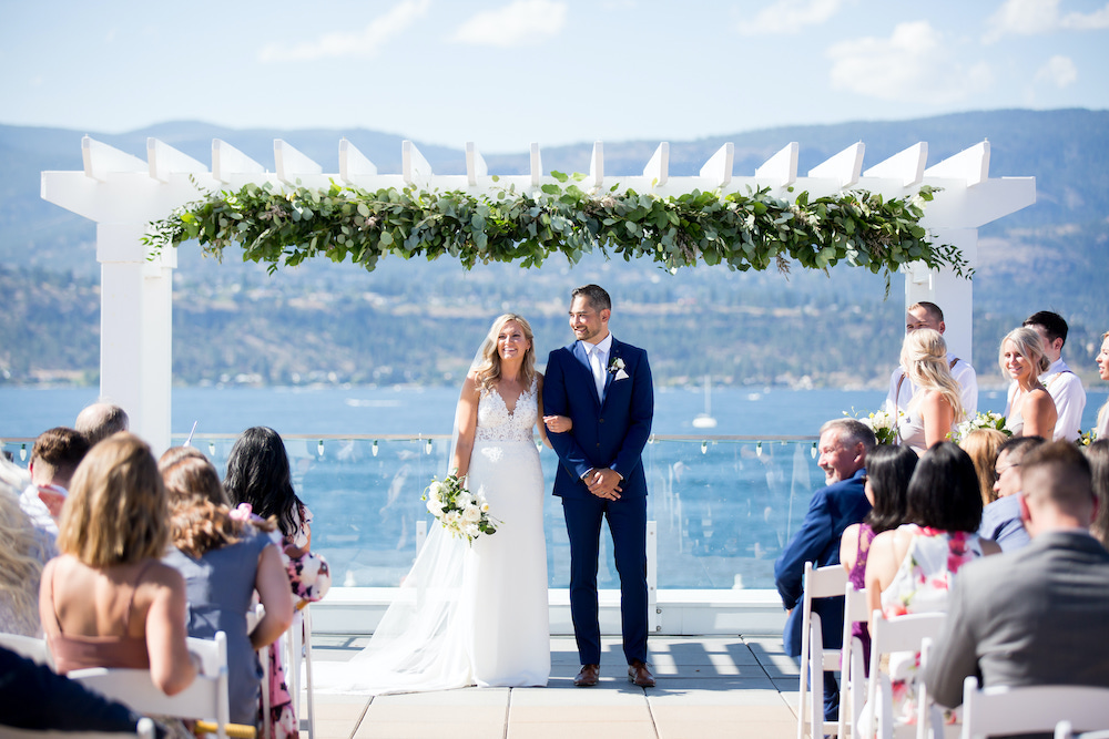 Wedding couple hold ceremony in the sunshine on lake pier with blue skies behind in Okanagan