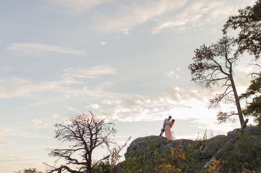 Engaged couple kiss at the top of hill with ocean sunset behind them