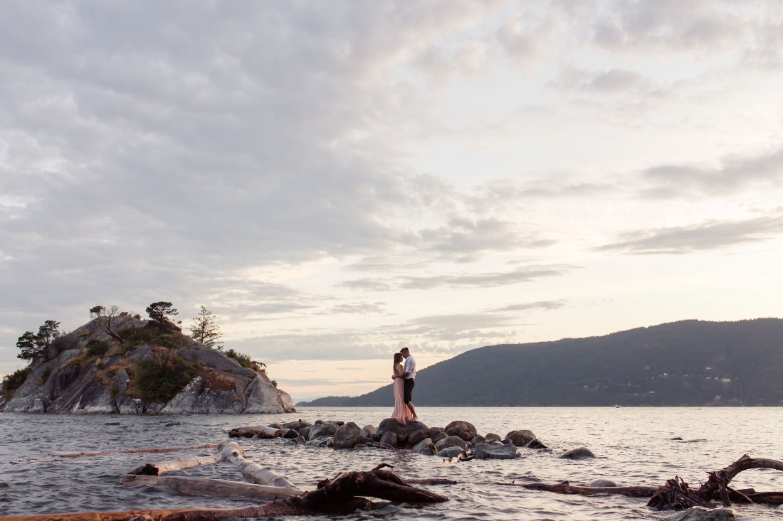 Couple in love pose on rocks in water with ocean sunset behind them in Vancouver