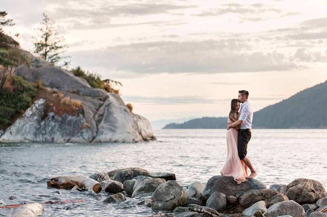 Engaged couple pose on beach rocks with ocean behind them