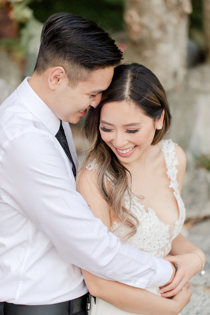 Bride to be wears long blush gown with lace top and smiles at fiance wearing white shirt and blue tie