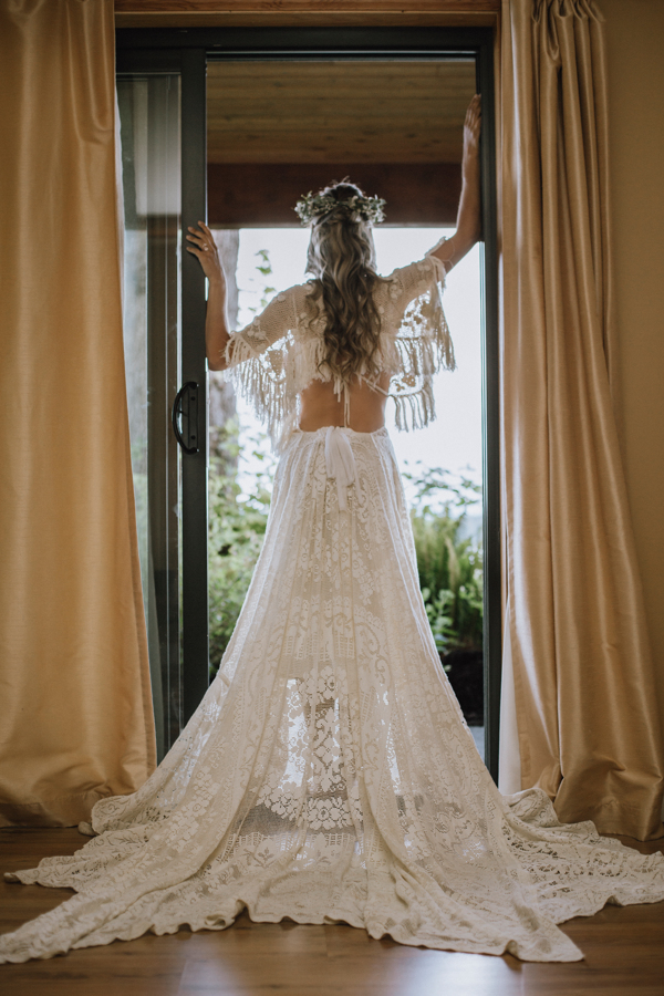 Bride looking out at ocean through window wearing stunning lace low back and flowing sleeves by Reclamation Design