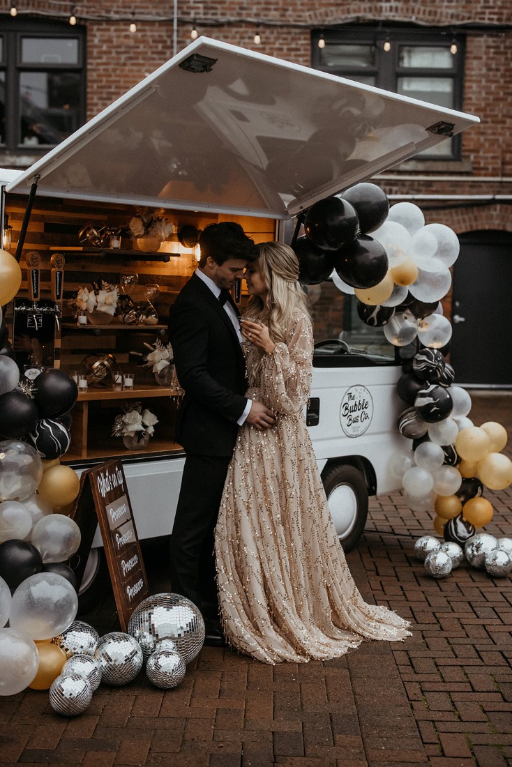 Glamorous couple enjoy personal moment sharing champagne in front of balloon garlands and bubble bus