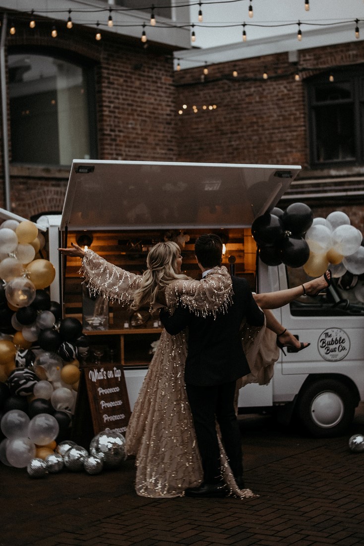 Couple twirl around drinking champagne in front of bubble bus co on Vancouver Island