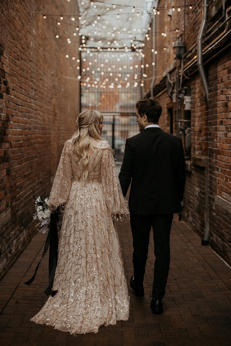 Romantic couple stroll through Vancouver Island alley wearing blush flitter gown and black suit with bowtie 