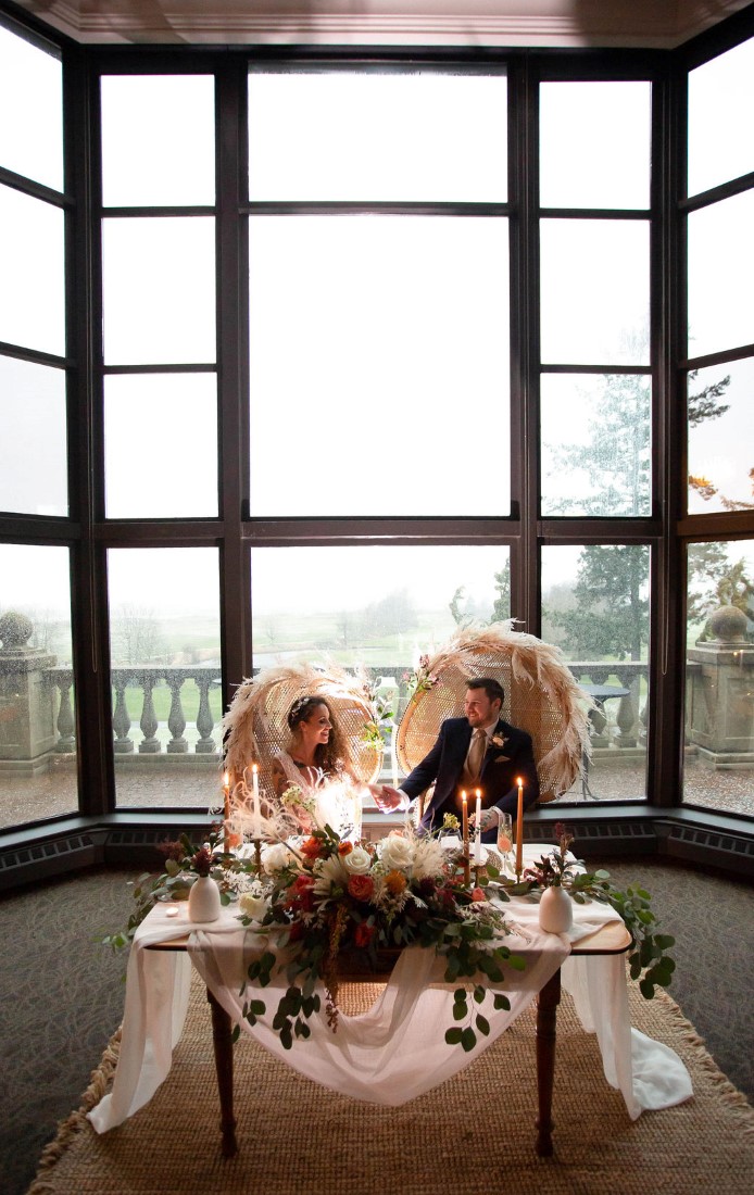 Bohemian Inspired Elopement couple sit in wicker chairs in front of a wall of windows at Swaneset Vancouver