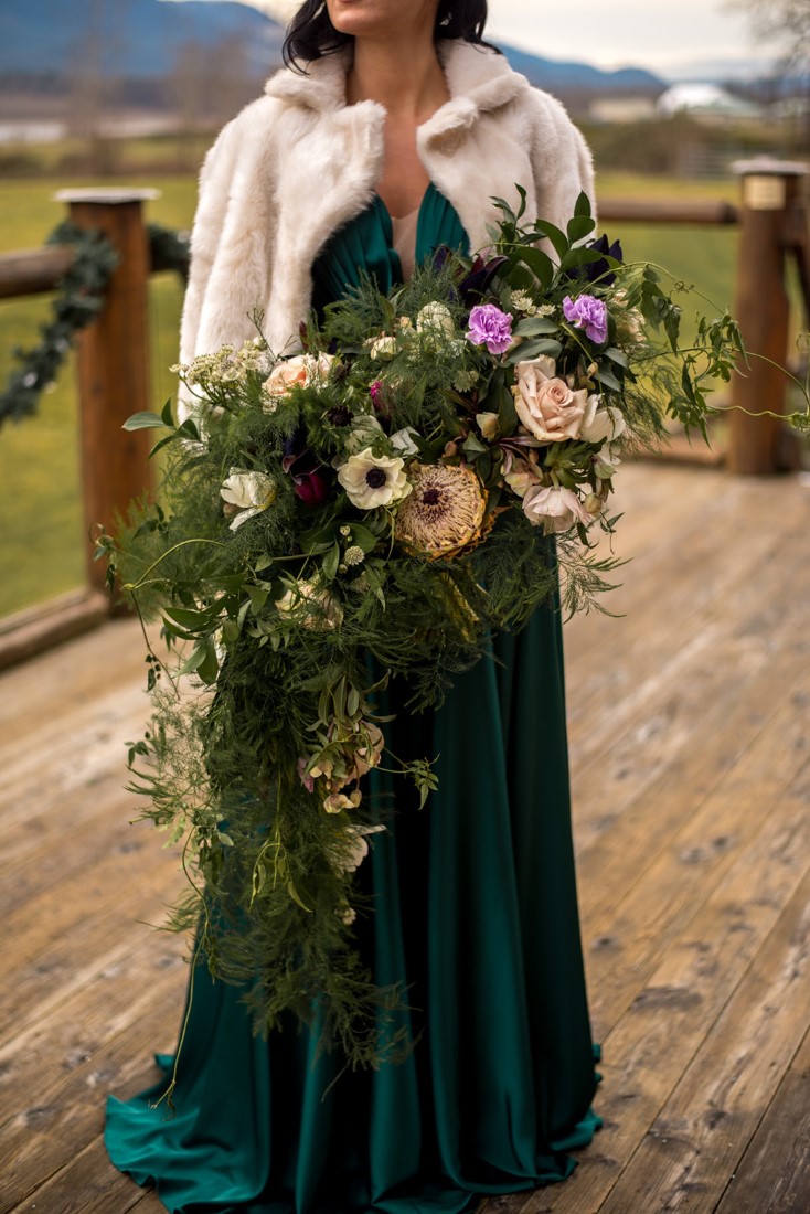 Bride in green gown holds bouquet of anemones, roses and greenery by All Her Flowers Fraser Valley