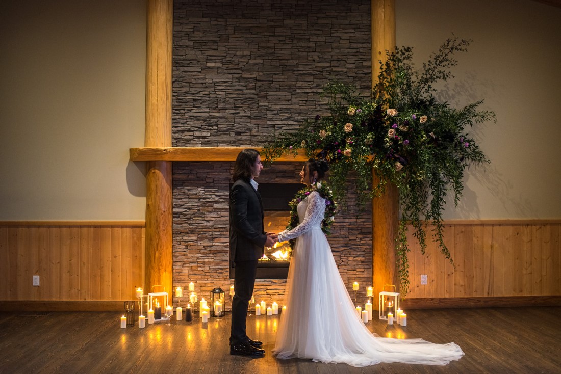 Newlyweds stand in front of large floral arrangement on fireplace mantle