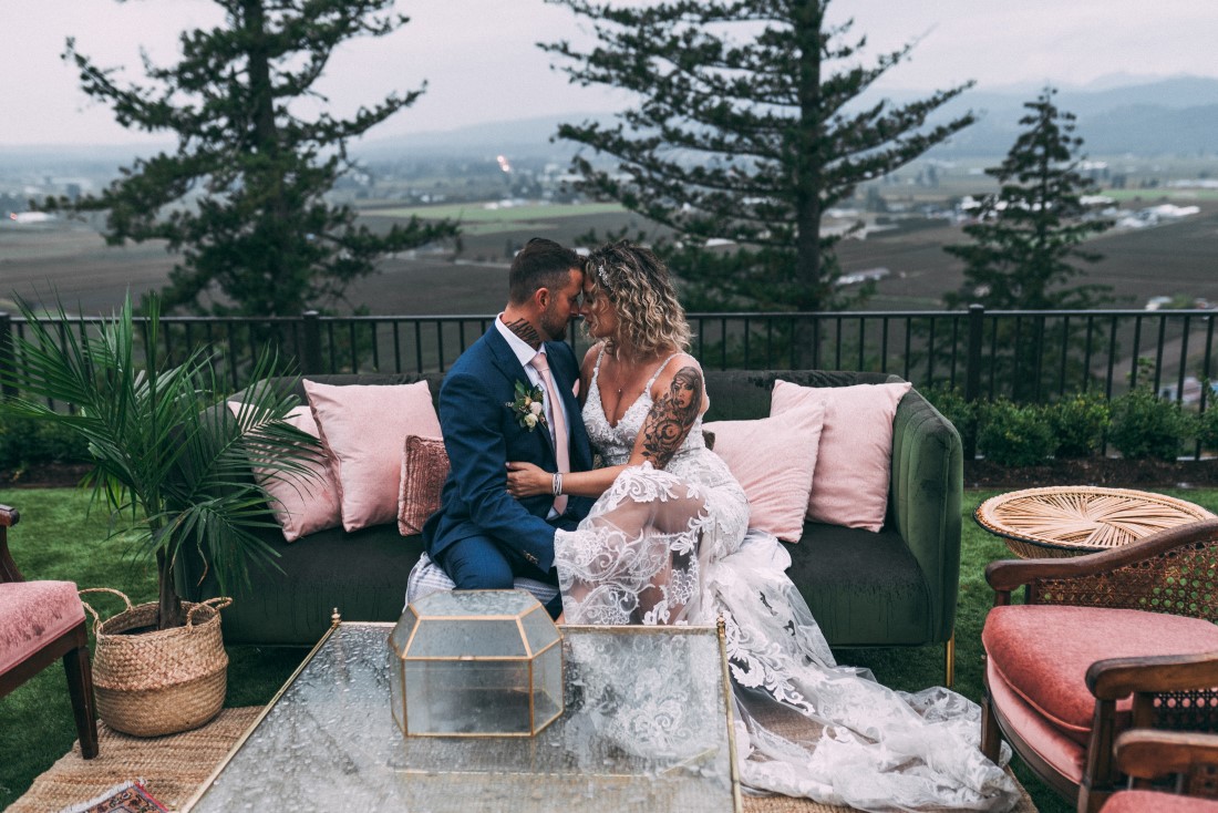 Newlyweds sit on couch with trees and mountains in the background by Vancouver