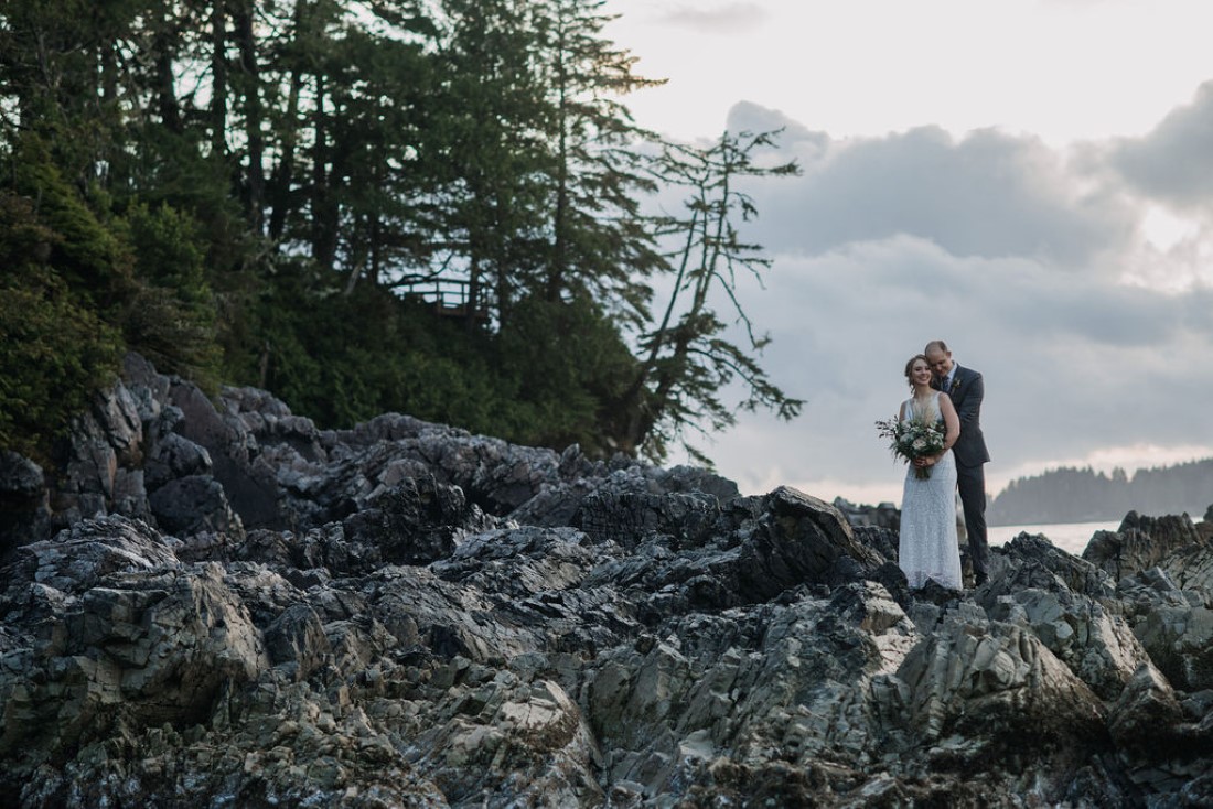 Newlyweds stand on black rock with trees and ocean behind them in Tofino