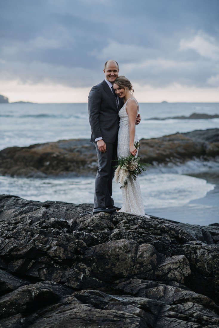 Newlyweds stand on black rocks of Ucluelet by Bracey Photography