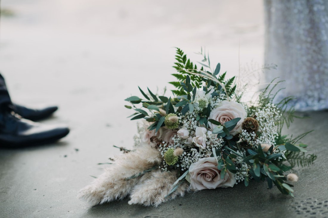 Bouquet of ferns, roses and dried grasses on beach sand by Crab Apple Floral Tofino